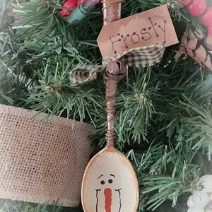 May include: A wooden spoon painted to look like a snowman with a red nose and black eyes. The spoon is hanging on a Christmas tree with a brown tag that says "Frosty".