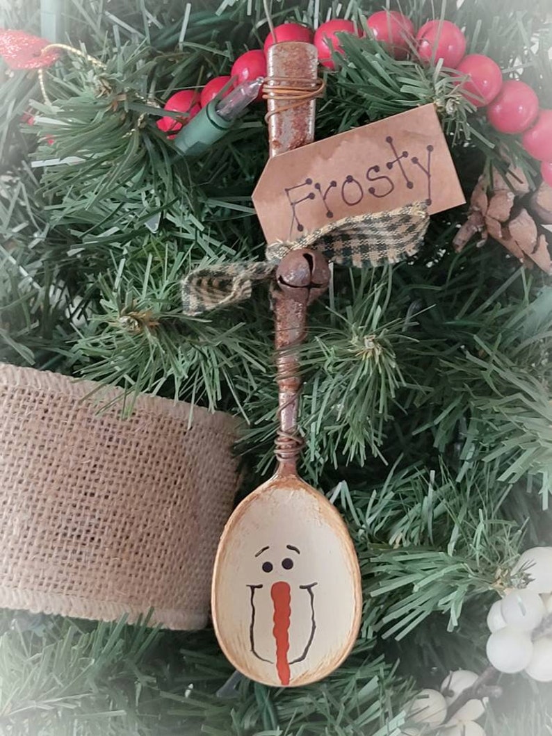 May include: A wooden spoon painted to look like a snowman with a red nose and black eyes. The spoon is hanging on a Christmas tree with a brown tag that says "Frosty".