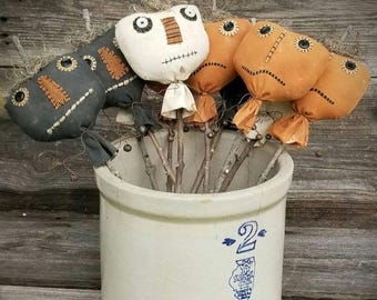 May include: A collection of fabric-covered, stuffed head shapes on sticks, arranged in a cream-coloured ceramic crock with the number 2 and a blue logo printed on the front. The heads feature button eyes and stitched mouths in black, white, and orange.