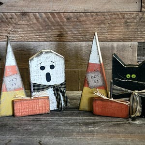 May include: A set of four wooden Halloween decorations including two triangular candy corn shapes with Oct 31 labels, a white ghost with a black and white chequered ribbon, a black cat with green eyes, and two small orange rectangular blocks.
