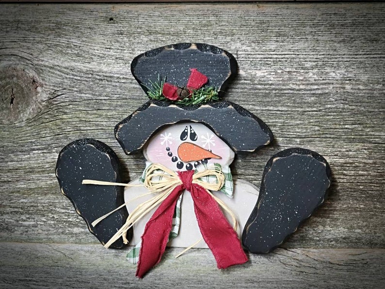 May include: A wooden snowman ornament with a black top hat, red scarf, and white face. The snowman has black arms and a white body with a green and white plaid pattern.