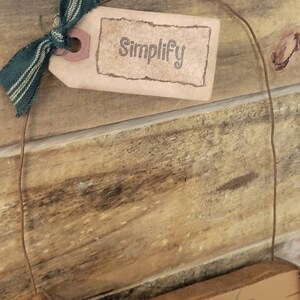 Puede incluir: Un letrero de madera r&uacute;stico con la palabra "Simplify" impresa en una etiqueta marr&oacute;n, atada con una cinta a rayas verdes y blancas. El letrero cuelga de una fina cuerda marr&oacute;n, sobre un fondo de madera.