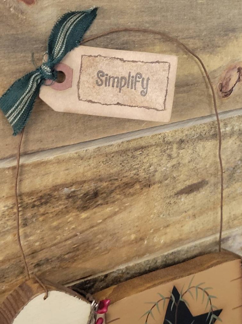 Puede incluir: Un letrero de madera r&uacute;stico con la palabra "Simplify" impresa en una etiqueta marr&oacute;n, atada con una cinta a rayas verdes y blancas. El letrero cuelga de una fina cuerda marr&oacute;n, sobre un fondo de madera.