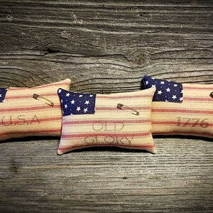 May include: Three small decorative pillows with a red, white, and blue striped fabric. Each pillow has a blue star patch on one side and the words "USA", "Old Glory", and "1776" on the other side.