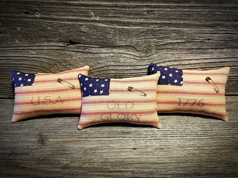 May include: Three small decorative pillows with a red, white, and blue striped fabric. Each pillow has a blue star patch on one side and the words "USA", "Old Glory", and "1776" on the other side.