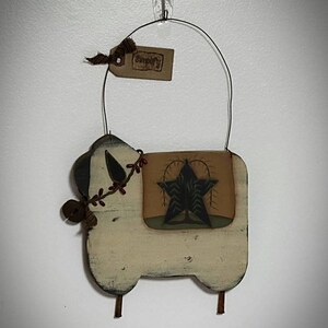 Puede incluir: Un colgante de pared r&uacute;stico de madera en forma de oveja, color crema, con acabado envejecido. Presenta un dise&ntilde;o de estrella, una etiqueta que dice "Simplify" y un asa de alambre. Acentuado con una peque&ntilde;a campana y vegetaci&oacute;n artificial. La oveja mide unos 25 cm de ancho.