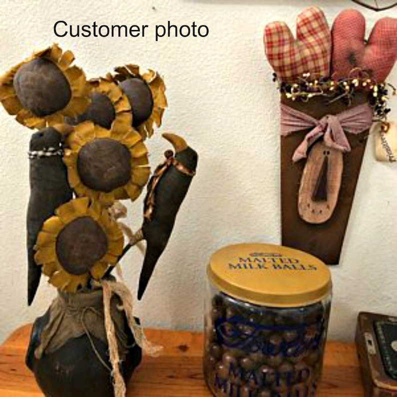 May include: A handmade primitive style sunflower and crow decoration with a black vase. The sunflowers are made of fabric and have a yellow and brown color scheme. The crows are black and have a rustic look. The vase is black and has a burlap bow around the top. The decoration is sitting on a wooden table next to a jar of chocolate candies.
