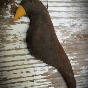May include: A black felt bird with a yellow beak hanging from a black string. The bird is a primitive folk art style and has a rustic look.