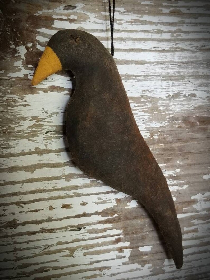 May include: A black felt bird with a yellow beak hanging from a black string. The bird is a primitive folk art style and has a rustic look.