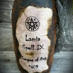 Op de afbeelding: Een vintage ogende fles met een etiket waarop staat "Lamia Spell IX Tongue of Dog 1609". Het etiket heeft een pentagram symbool en is lichtjes verbrand rond de randen. De fles is gewikkeld in een stuk gescheurd stof.