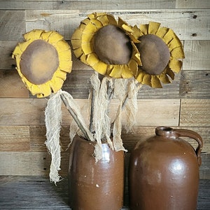 May include: Three yellow felt sunflowers with brown centers in a brown ceramic vase. The vase is sitting on a wooden surface.
