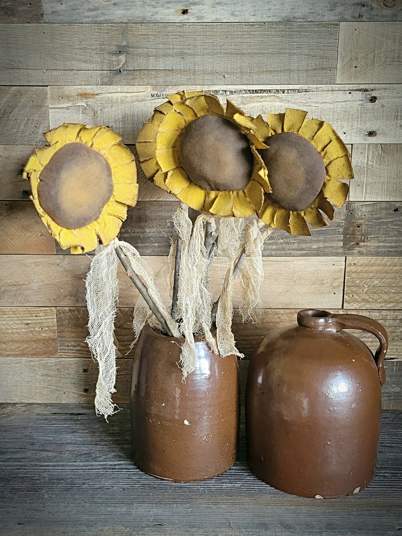 May include: Three yellow felt sunflowers with brown centers in a brown ceramic vase. The vase is sitting on a wooden surface.