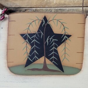 Puede incluir: Un letrero de madera r&uacute;stico y rectangular con una estrella negra con un dise&ntilde;o de sauce llor&oacute;n. La estrella est&aacute; centrada en un fondo beige con una base verde, y el letrero tiene un colgador de alambre.