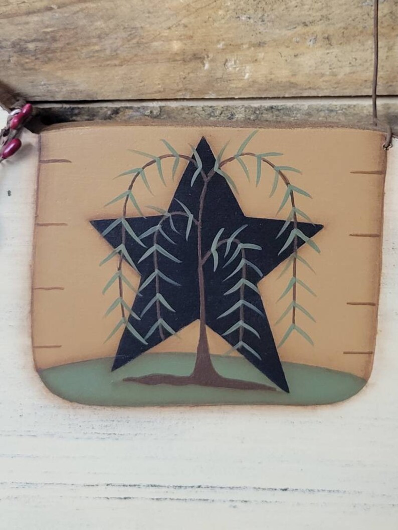 Puede incluir: Un letrero de madera r&uacute;stico y rectangular con una estrella negra con un dise&ntilde;o de sauce llor&oacute;n. La estrella est&aacute; centrada en un fondo beige con una base verde, y el letrero tiene un colgador de alambre.