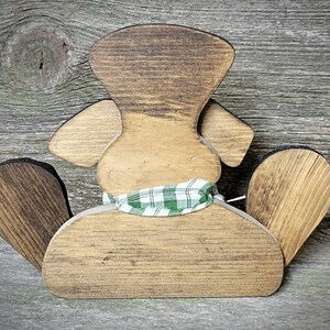 May include: Wooden cutout of a dog wearing a green and white plaid scarf. The dog is sitting with its back to the viewer. The dog is made of unfinished wood and has a rustic look.