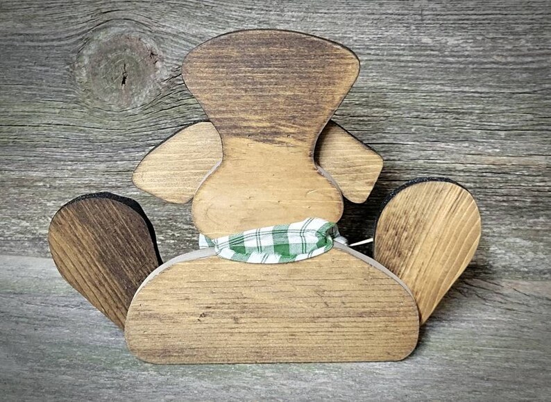 May include: Wooden cutout of a dog wearing a green and white plaid scarf. The dog is sitting with its back to the viewer. The dog is made of unfinished wood and has a rustic look.
