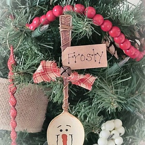 May include: A spoon ornament painted to look like a snowman with a red nose and black eyes. The spoon is attached to a brown wire hanger with a red and white plaid ribbon and a small bell. The ornament is labeled "Frosty" on a brown tag.