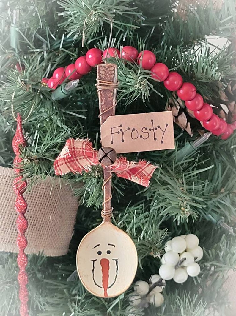 May include: A spoon ornament painted to look like a snowman with a red nose and black eyes. The spoon is attached to a brown wire hanger with a red and white plaid ribbon and a small bell. The ornament is labeled "Frosty" on a brown tag.