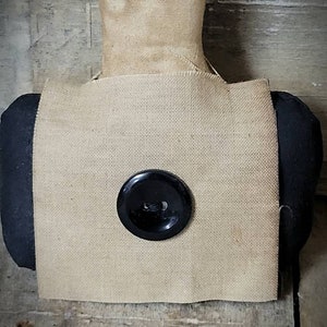 May include: A square of brown burlap fabric with a black button in the centre. The fabric is attached to a black fabric covered form.