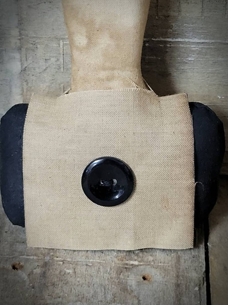 May include: A square of brown burlap fabric with a black button in the centre. The fabric is attached to a black fabric covered form.