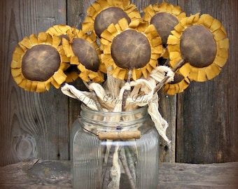 May include: A collection of fabric sunflowers with dark brown centers and mustard yellow petals, tied together with frayed cream-colored fabric strips, displayed in a clear, vertically ribbed glass jar with a metal wire handle.