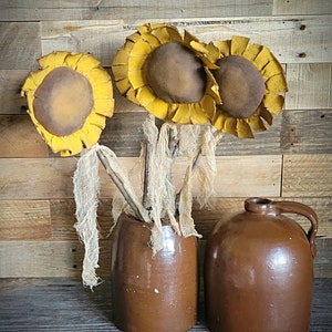 May include: Three brown fabric sunflowers with yellow petals in a brown ceramic vase and a brown ceramic jug.