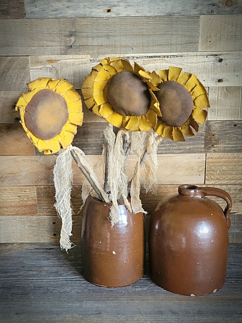 May include: Three brown fabric sunflowers with yellow petals in a brown ceramic vase and a brown ceramic jug.