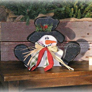 May include: A wooden snowman figurine with a black hat, a white face, a red scarf, and a plaid bow. The snowman is standing on a wooden shelf with a sign that says "Let it Snow".