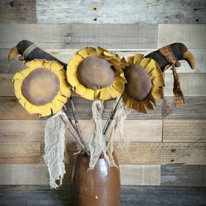 May include: Three brown and yellow fabric sunflowers with brown bird heads and plaid ribbon accents in a brown ceramic vase.
