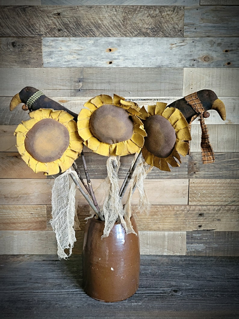 May include: Three brown and yellow fabric sunflowers with brown bird heads and plaid ribbon accents in a brown ceramic vase.
