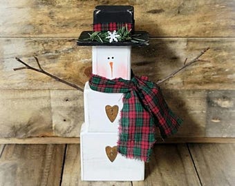 Puede incluir: Una figura de muñeco de nieve hecha de tres bloques de madera blancos apilados. Presenta un sombrero de copa negro con una banda de cuadros rojos y verdes, una bufanda de tela a cuadros a juego, dos botones marrones en forma de corazón y brazos de ramitas.