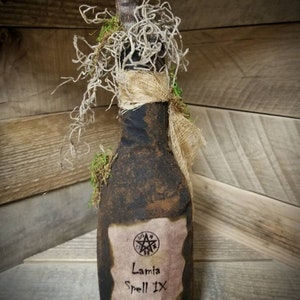 Op de afbeelding: Een donkerbruine glazen fles met een kurkstop en een etiket met de tekst "Lamia Spell IX Tongue of Dog 1609". De fles is bedekt met mos en touw.