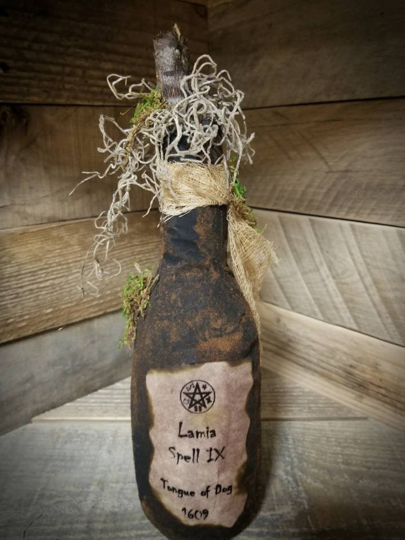 Op de afbeelding: Een donkerbruine glazen fles met een kurkstop en een etiket met de tekst "Lamia Spell IX Tongue of Dog 1609". De fles is bedekt met mos en touw.