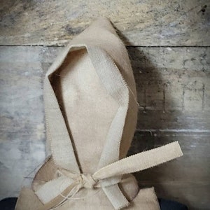 May include: A handmade doll with a beige fabric hood tied at the neck. The doll is wearing a beige fabric dress and has a dark fabric body.