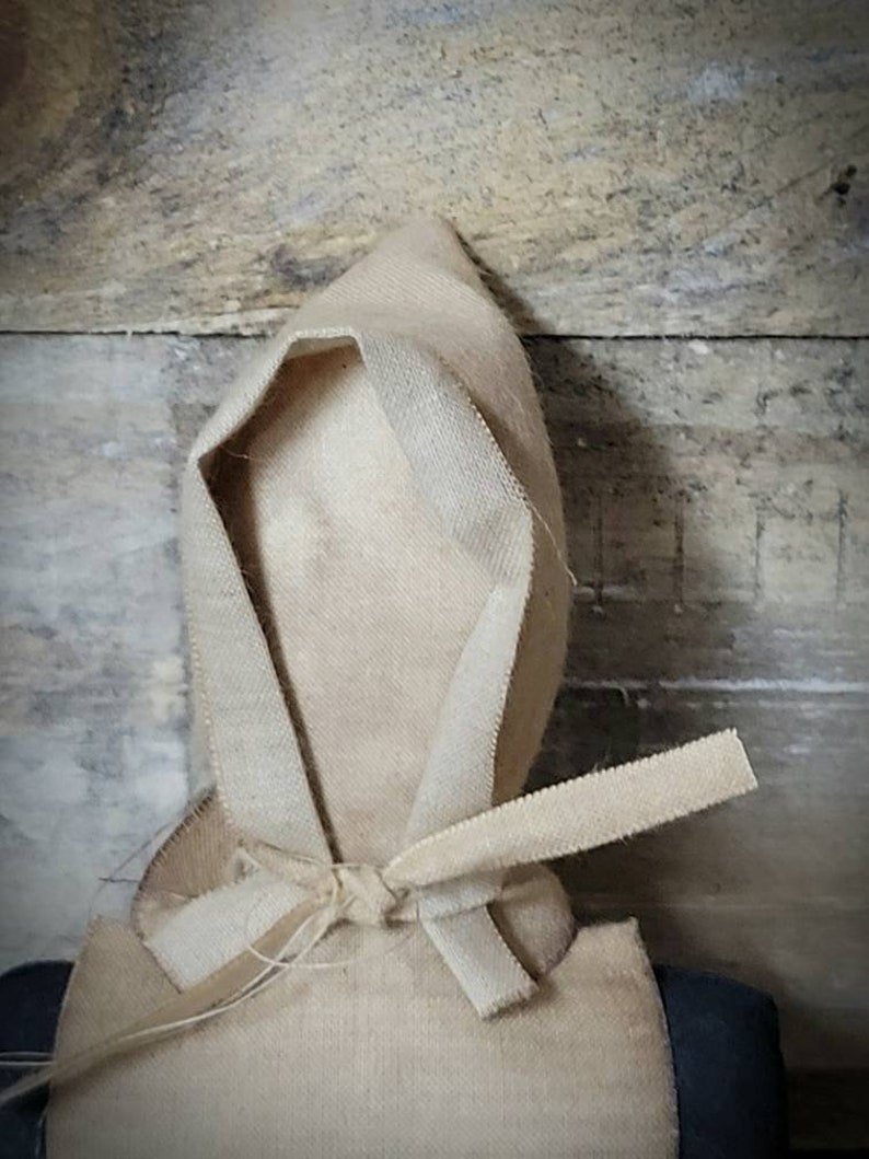 May include: A handmade doll with a beige fabric hood tied at the neck. The doll is wearing a beige fabric dress and has a dark fabric body.