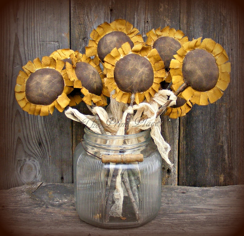 May include: A bouquet of brown and yellow fabric sunflowers in a clear glass jar with a wooden handle. The sunflowers are arranged in a vase with burlap ribbon and twigs.