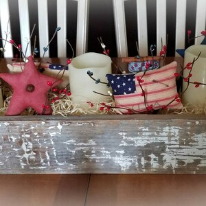 May include: A rustic wooden box centerpiece decorated with red and white berry sprigs, three white pillar candles, a red fabric star, and a red and white striped pillow with a blue star.