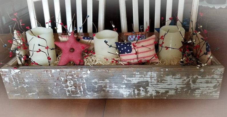 May include: A rustic wooden box centerpiece decorated with red and white berry sprigs, three white pillar candles, a red fabric star, and a red and white striped pillow with a blue star.