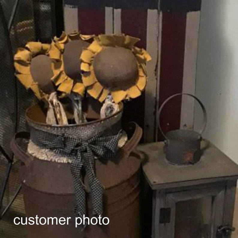 May include: A rustic milk can with a burlap and black and white checkered ribbon bow, decorated with three yellow felt sunflowers. The milk can is sitting on a wooden surface next to a small black metal lantern.