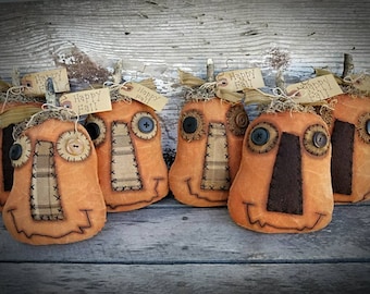 May include: Several orange fabric pumpkins with stitched features, button eyes, and plaid patches. Each pumpkin has a twig stem, raffia, and a tag saying "Happy Fall!" on a rustic wooden surface.