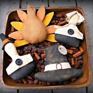 May include: A wooden bowl filled with cinnamon sticks and potpourri, with four handmade Thanksgiving-themed fabric figures: a turkey, a pilgrim, and two pilgrims wearing hats. The text "The Prim Place" is visible on the bowl.