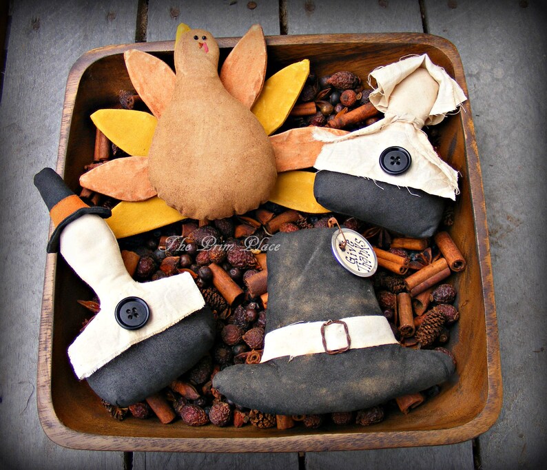 May include: A wooden bowl filled with cinnamon sticks and potpourri, with four handmade Thanksgiving-themed fabric figures: a turkey, a pilgrim, and two pilgrims wearing hats. The text "The Prim Place" is visible on the bowl.