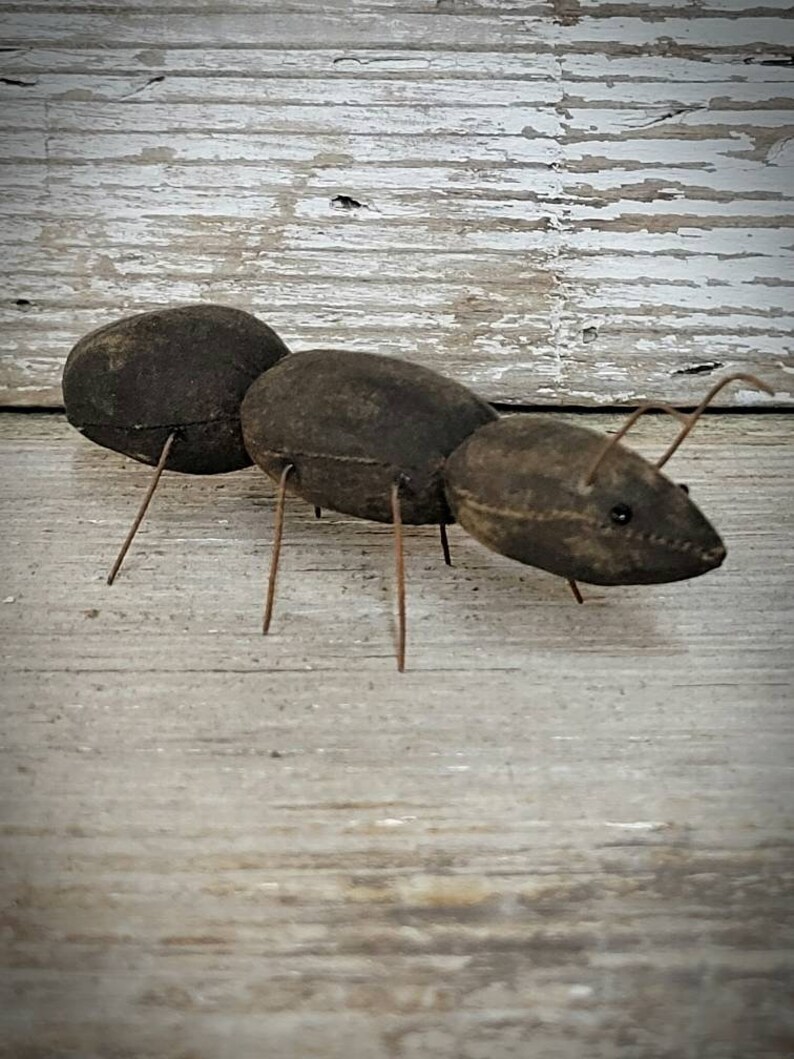 Pu&ograve; includere: Una formica di stoffa marrone con gambe di filo metallico. La formica &egrave; seduta su una superficie di legno marrone chiaro.
