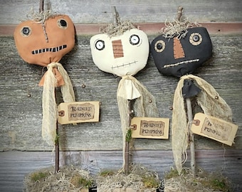 May include: Three fabric pumpkin figures in orange, white, and black, each with button eyes, stitched mouths, and twig stems. They feature frayed fabric neck wraps, mossy wooden bases, and tan tags with the text Haunted Pumpkins.