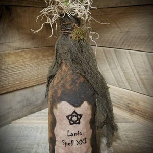 Op de afbeelding: Een bruine glazen fles met een etiket waarop staat "Lamia Spell XXX Eye of Newt 1609". De fles is gewikkeld in jute en heeft een met mos bedekte bovenkant.