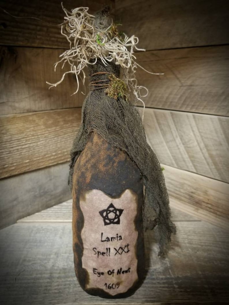 Op de afbeelding: Een bruine glazen fles met een etiket waarop staat "Lamia Spell XXX Eye of Newt 1609". De fles is gewikkeld in jute en heeft een met mos bedekte bovenkant.