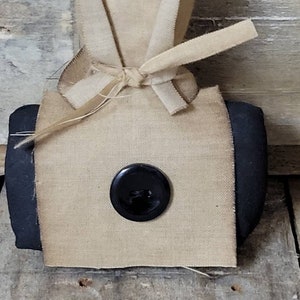 May include: A small, square-shaped, beige fabric bag with black trim. The bag is tied with a beige ribbon and has a black button in the centre.