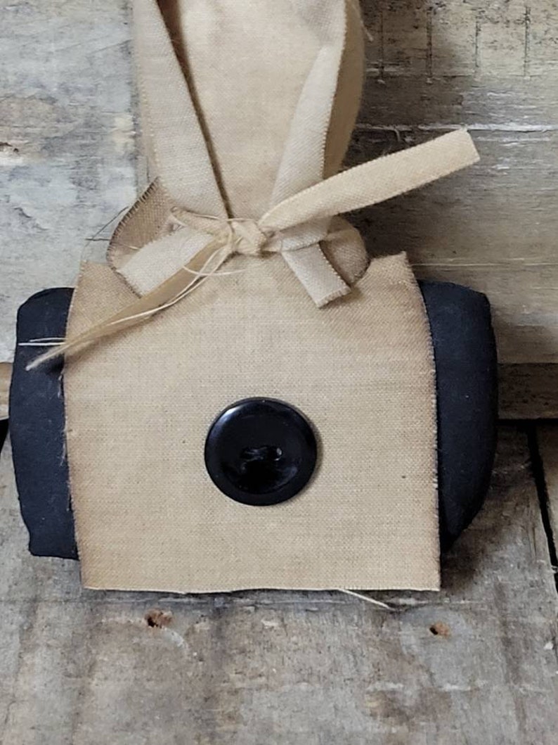 May include: A small, square-shaped, beige fabric bag with black trim. The bag is tied with a beige ribbon and has a black button in the centre.