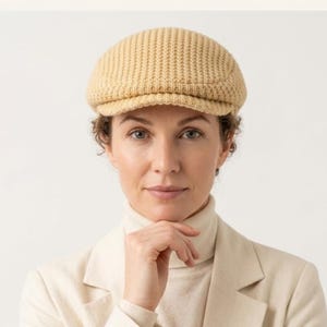 May include: A light beige flat cap featuring a textured, waffle-knit pattern. The cap has a rounded crown and a short, stiff brim extending from the front. The material appears to be a soft, woven knit fabric with a consistent, uniform color throughout.