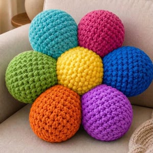 May include: A flower-shaped cushion made of seven interconnected crocheted spheres in bright colors. The center sphere is yellow, surrounded by six spheres in turquoise, pink, blue, purple, orange, and light green, all featuring a textured yarn stitch.